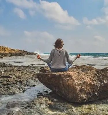 Femme assise sur un rocher face à la mer, mains écartées, doigts joints en posture de méditation