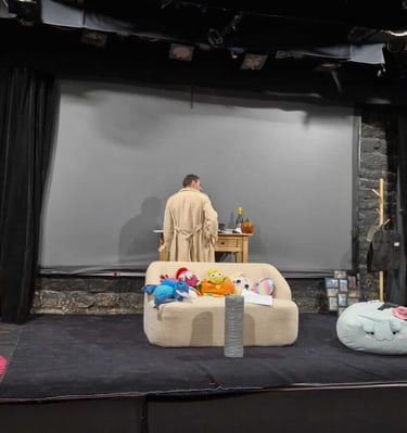 Scène de théâtre en décor domestique : une personne en robe beige est debout à une table, entourée d