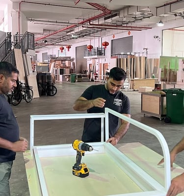 Professional carpenters assembling a white custom furniture frame using a power drill in a workshop.