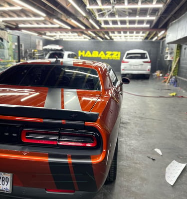 Orange Dodge Challenger with black racing stripes undergoing custom vinyl wrap at Habazzi Auto Styles garage.