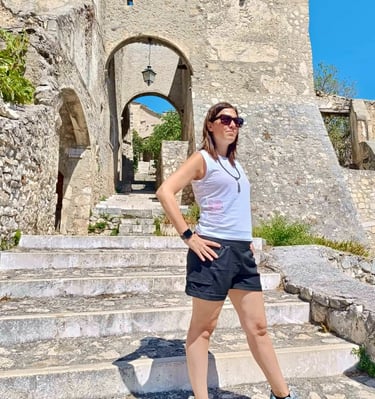 A female traveler in sunglasses stands on stone steps at an ancient European fortress gateway.