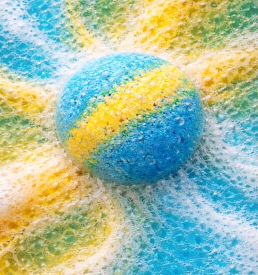 Oversized bath bomb releasing blue and yellow foam in soft, cloud-like fizz.