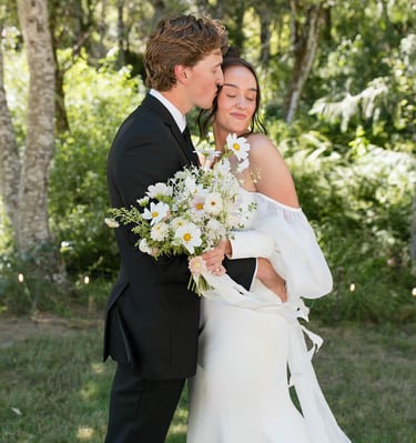 Groom in black tuxedo kissing bride in off-the-shoulder white wedding dress holding a daisy bouquet.