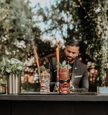 Professional bartender serving tropical tiki cocktails with sparkling garnishes at an outdoor garden bar.