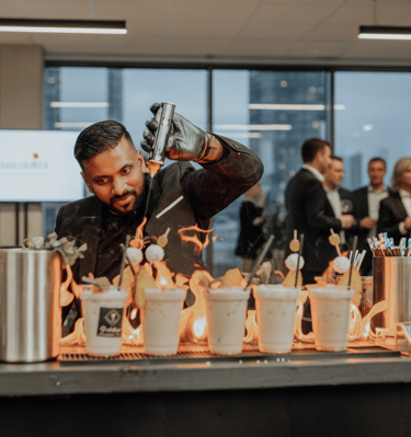 A professional bartender uses a torch to create a fire display over tropical cocktails at a corporate event.