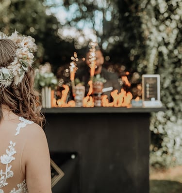 A bride with a floral crown watches a flair bartender perform a fire show at an outdoor wedding reception.