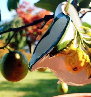 A decorative ceramic lemon creamer pitcher hanging from a green lemon tree branch outdoors.