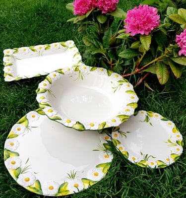 Hand-painted ceramic dinnerware set with 3D daisy flowers displayed on green grass by pink rhododendrons.
