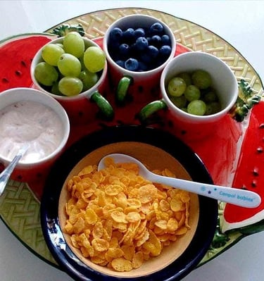 Healthy breakfast tray with cornflakes, yogurt, fresh grapes, and blueberries in a watermelon dish.