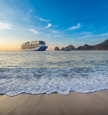 A large luxury cruise ship anchored in the ocean near a tropical beach and rocky cliffs at sunset.