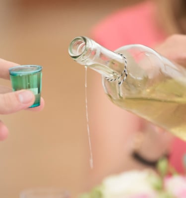 A hand pouring a clear alcoholic beverage from a glass bottle into a small blue shot glass.