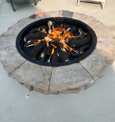 A round outdoor stone gas fire pit with burning ceramic logs on a concrete patio.