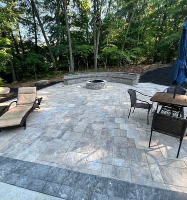 A backyard stone paver patio featuring a fire pit, curved stone bench, and outdoor dining set with lounge chairs.