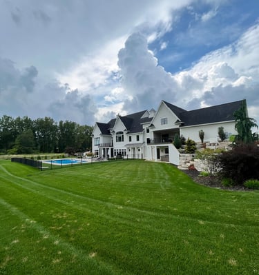 Modern white farmhouse mansion with a luxury backyard pool and manicured green lawn under a cloudy sky.