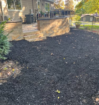 Backyard patio with stone retaining wall and fresh black mulch landscaping in a residential garden.