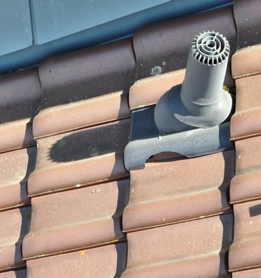 A grey plastic roof vent pipe installed on brown terracotta clay tiles for attic ventilation.