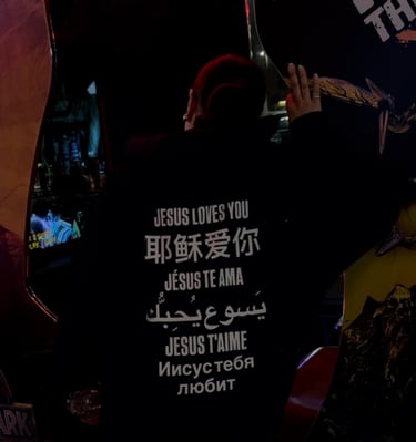 Back view of person in hoodie with 'Jesus loves you' in many languages, arms raised in neon arcade.