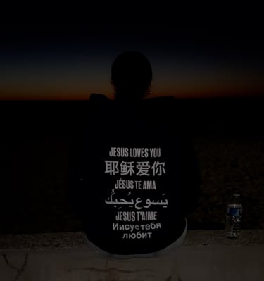 Silhouetted person at sunset wearing hoodie reads Jesus loves you in many languages worldwide faith!