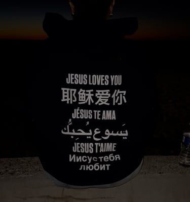 Silhouetted person at sunset wearing a hoodie that reads Jesus loves you in many languages.