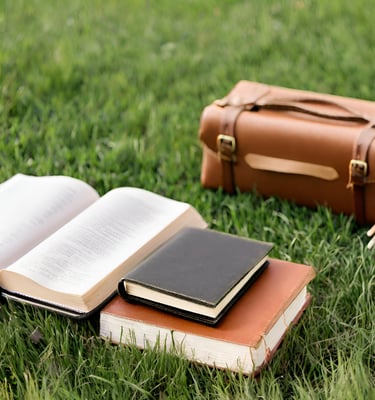 A satchel, two closed books, and one open book laying on a field of green grass.
