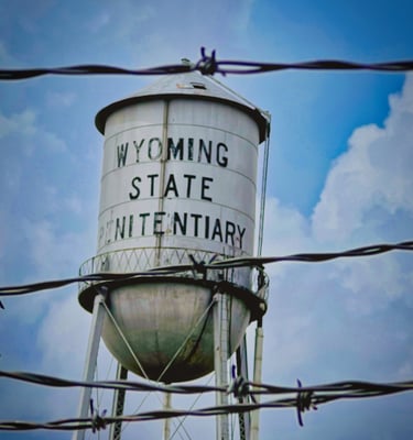 The Wyoming State Penitentiary water tower viewed through prison barbed wire under a cloudy sky.