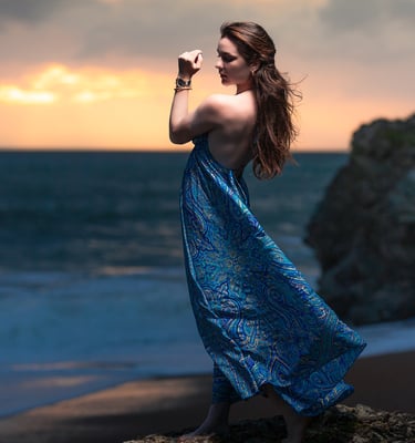 Creative portrait of a woman in a flowing blue dress standing on coastal rocks at sunset.