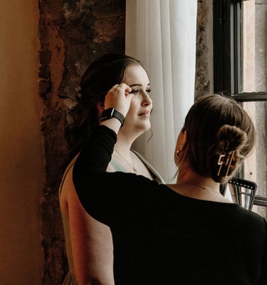Professional makeup artist applying false eyelashes to a bride by a window.