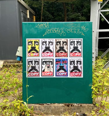 Urban street art stickers and graffiti tags on a green utility box in a park setting.