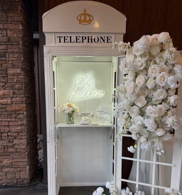 Vintage white telephone booth with floral arrangements and neon sign for wedding guestbook audio recording.