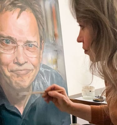 Stefanie placing final touches on oil portrait painting of smiling man with glasses.