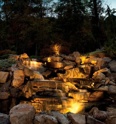 Illuminated backyard stone waterfall feature with landscape lighting and woodland backdrop.