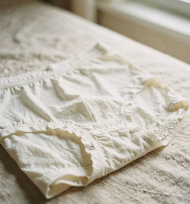 Soft film photography, close-up details of clean, folded organic cotton underwear on warm beige linen sheet, soft morning window light, 35mm.