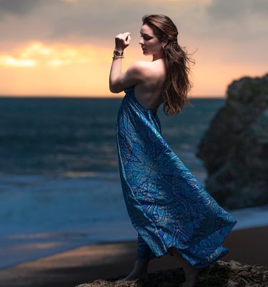 Portrait créatif d’une femme en robe bleue posant sur des rochers face à la mer au coucher du soleil