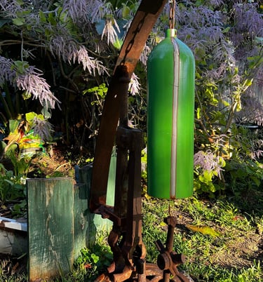 Rustic garden wind chime made from a repurposed green oxygen tank hanging from antique metal farm machinery.