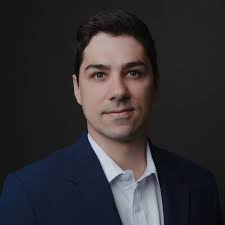 Homem vestindo terno azul escuro e camisa branca, posando diante de fundo preto.