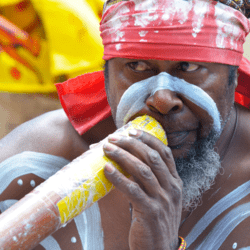 an Australian Aboriginal with white ochre on his face and long beard playing a didgeridoo