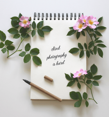 Spiral-bound notebook with pink dogwood flowers scattered about. "Stock photography is hard," written in black on the page.