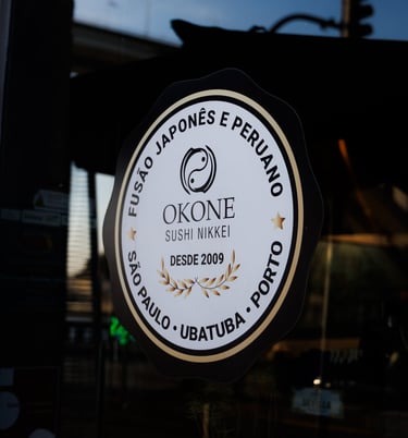Selo no vidro do Okone Sushi Nikkei com os dizeres Fusão Japonês e Peruano, citando São Paulo, Ubatuba e Porto desde 2009
