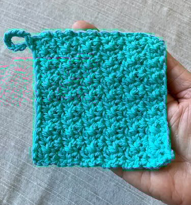 A hand holding a handmade crocheted aqua face scrubby with textured stitch