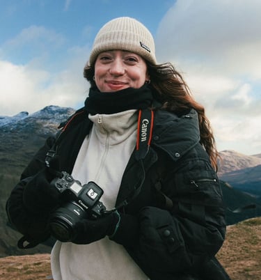 A photographer posing and smiling at the camera, holding her own camera, with mountain ranges in the background.  
