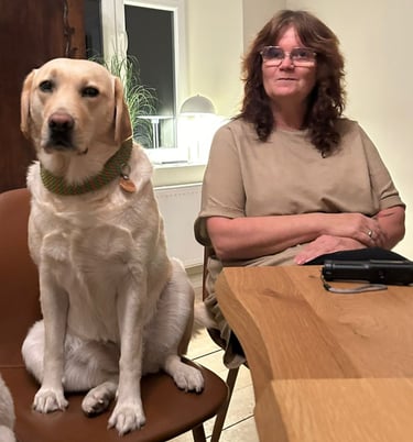 Carmen und ihre Labradorhündin Bonny sitzen an einem Tisch. Auch Bonny sitzt auf einem Stuhl.