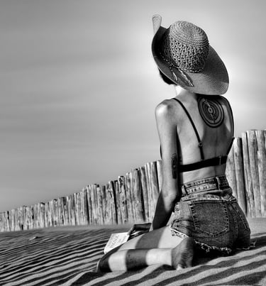 Photo artistique noire et blanc d'une jeune femme en mini short a genoux sur la plage avec un livre et un grand chapeau