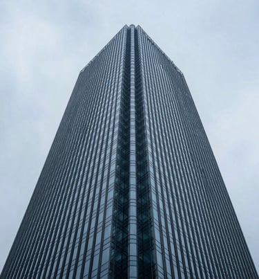 A low-angle shot of a minimalist skyscraper reaching into a light blue gray sky. Symmetrical composition, sleek glass reflections, Global / General urban setting.