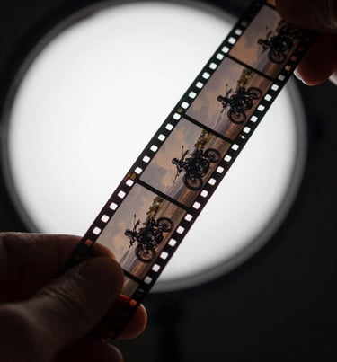 A close-up shot of a hand holding a strip of black and white film negatives up to a soft grey light source, revealing the frames of a motorcycle trip. Global / Western darkroom setting.