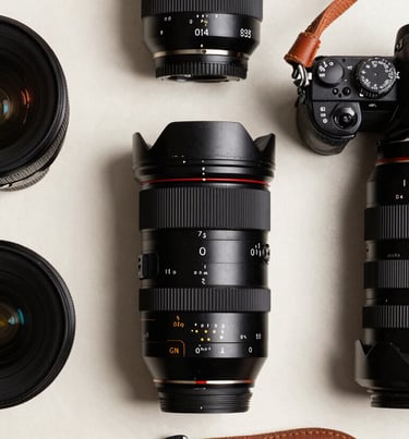 A high-contrast flat lay of professional photography equipment including lenses, a jet black camera body, and leather straps on an off-white background. Global / Western studio environment.