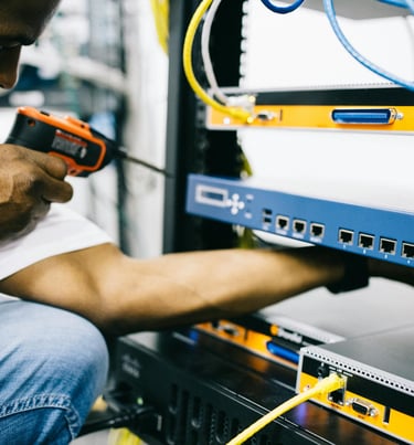 A close-up of a person working on a server rack, connecting network cables with a drill in hand and visible Ethernet ports.