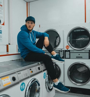 A man in a blue hoodie and cap sits casually on a washing machine in a laundromat. Several dryers are lined up in the backgro