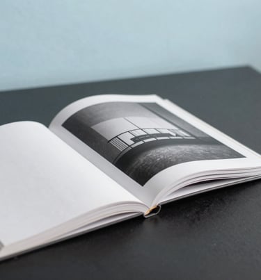 Close-up photography of a high-end photography book lying open on a dark charcoal table. The pages show minimalist black and white architecture. The background is a soft, out-of-focus baby blue wall.