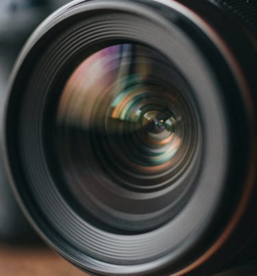 Artistic close-up of a lens reflection or camera detail in a dark, moody environment. Colors: #0D0D0D and subtle #6B705C highlights.