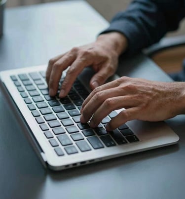 A close-up photograph of a North American / US person's hands typing on a sleek wireless keyboard on a clean steel blue desk.